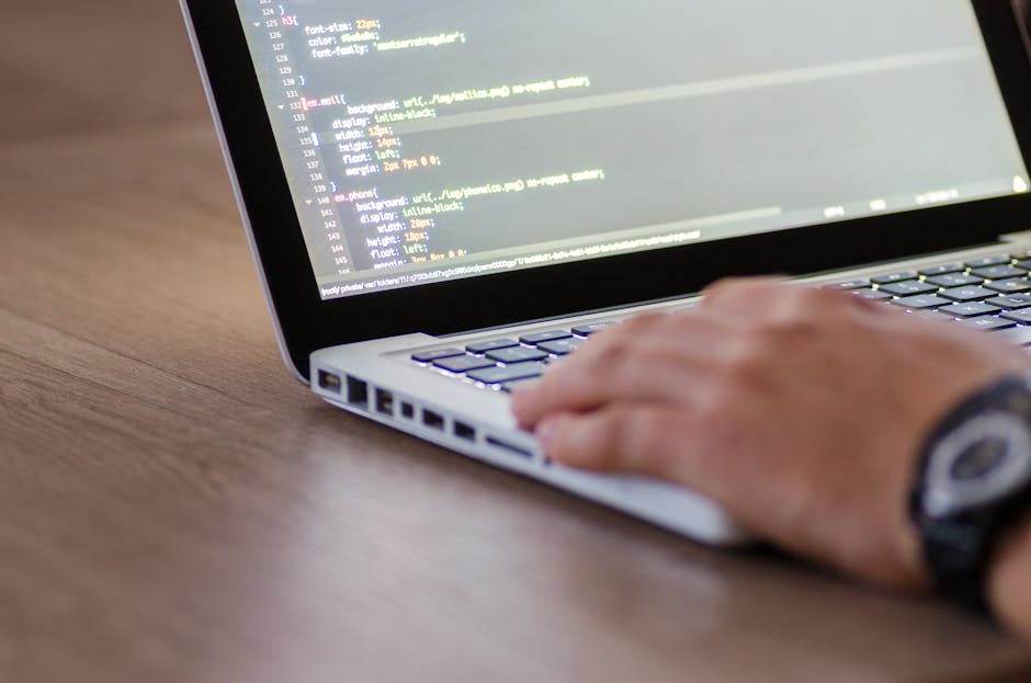 A close-up image of a person's hand typing on a silver laptop keyboard placed on a wooden surface. The laptop screen displays lines of visible programming code with a dark background and colorful synt